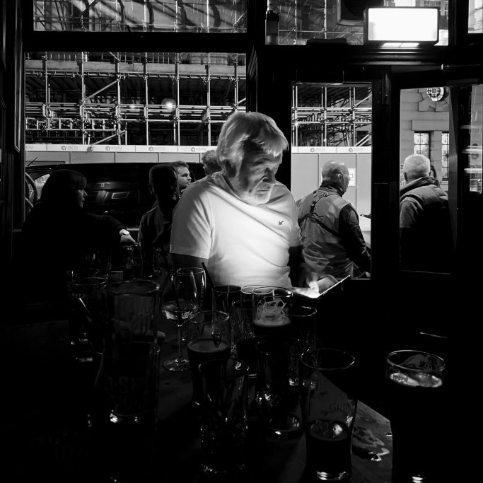 By the light of a silvery phone Black and white photograph of a man in pub looking at phone.